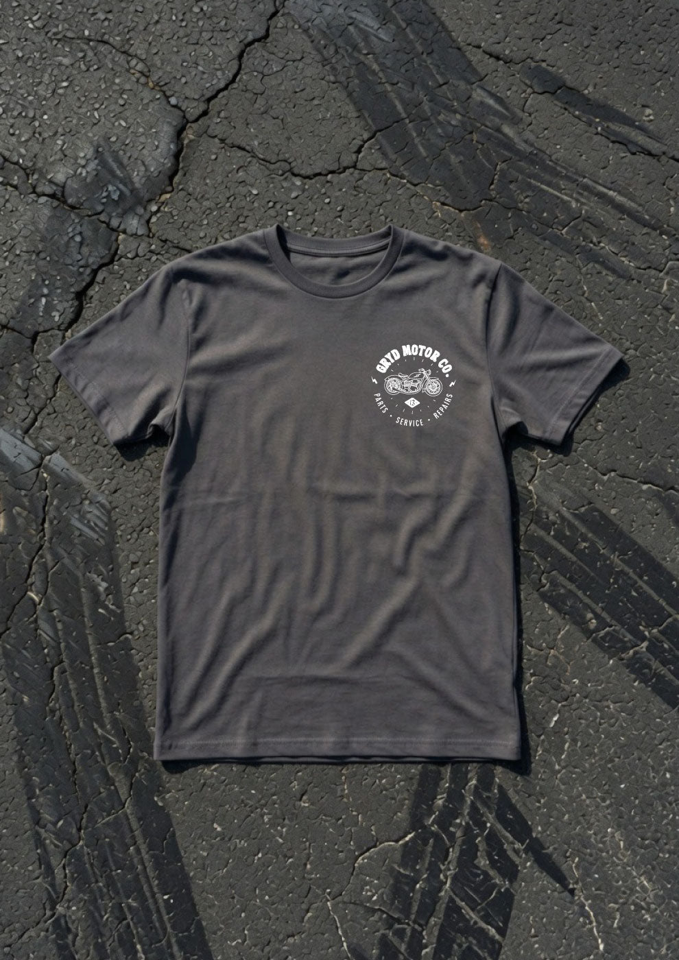 A dark charcoal grey t-shirt with a white circular graphic on the left chest, laid flat on a textured black asphalt surface with tire tracks.