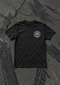 A black short-sleeve t-shirt with a circular white graphic on the left chest, laid flat on a textured dark asphalt surface.