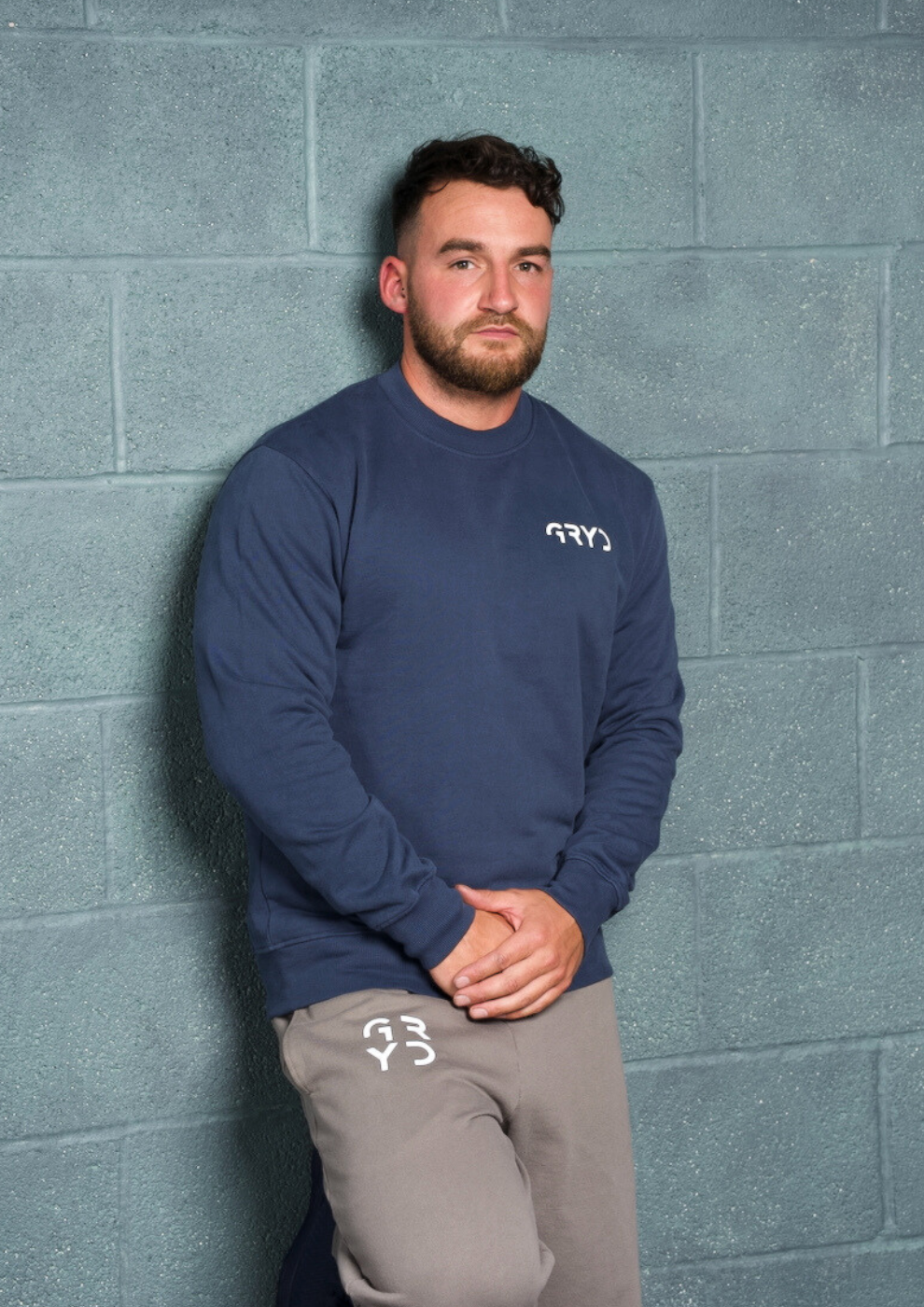 Man wearing a navy long-sleeve shirt and gray pants with a logo against a gray brick wall.