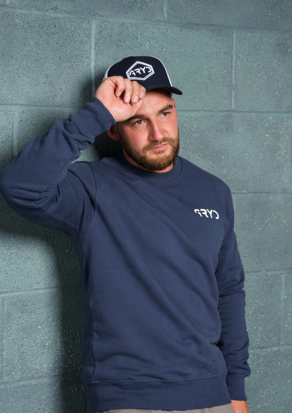Man wearing a navy blue sweatshirt and cap with a logo against a gray brick wall