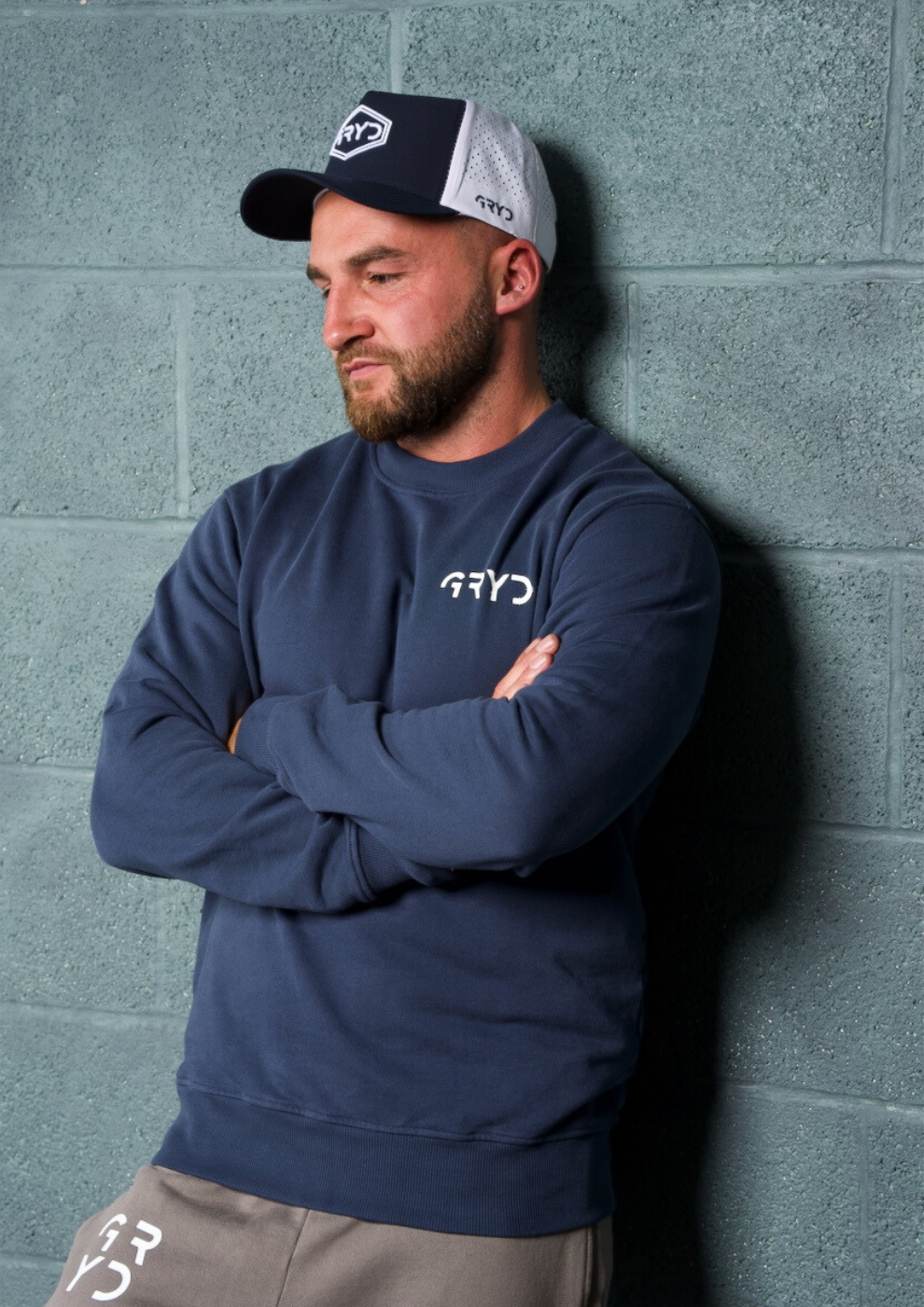 Man wearing a navy sweatshirt and cap with a logo, standing against a gray brick wall.
